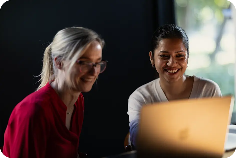 Two women smiling while discussing something on a laptop screen.
